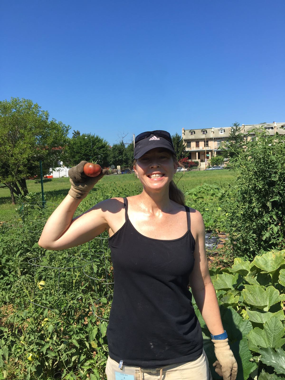Woman standing with a tomato in her hand in the midst of her community garden, with a big smile on her face.