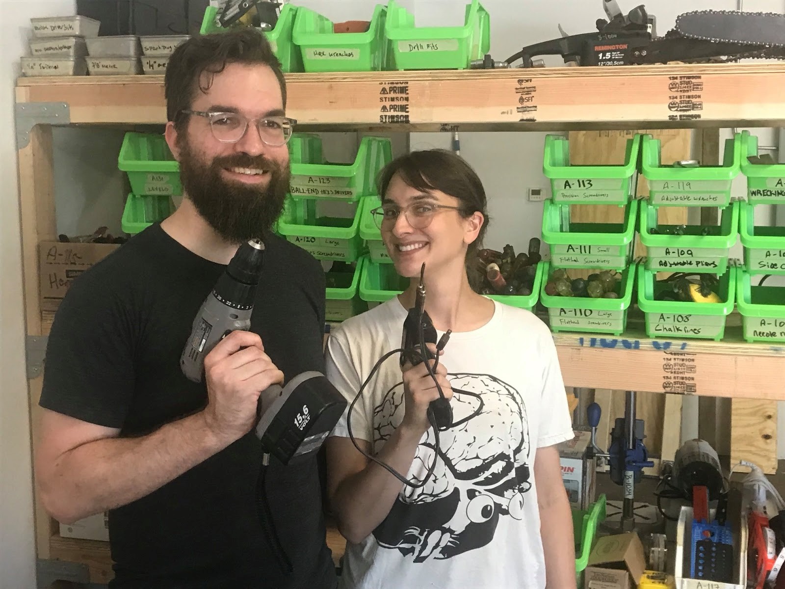 Smiling man and woman holding tools in front of shelves with more tools in bins