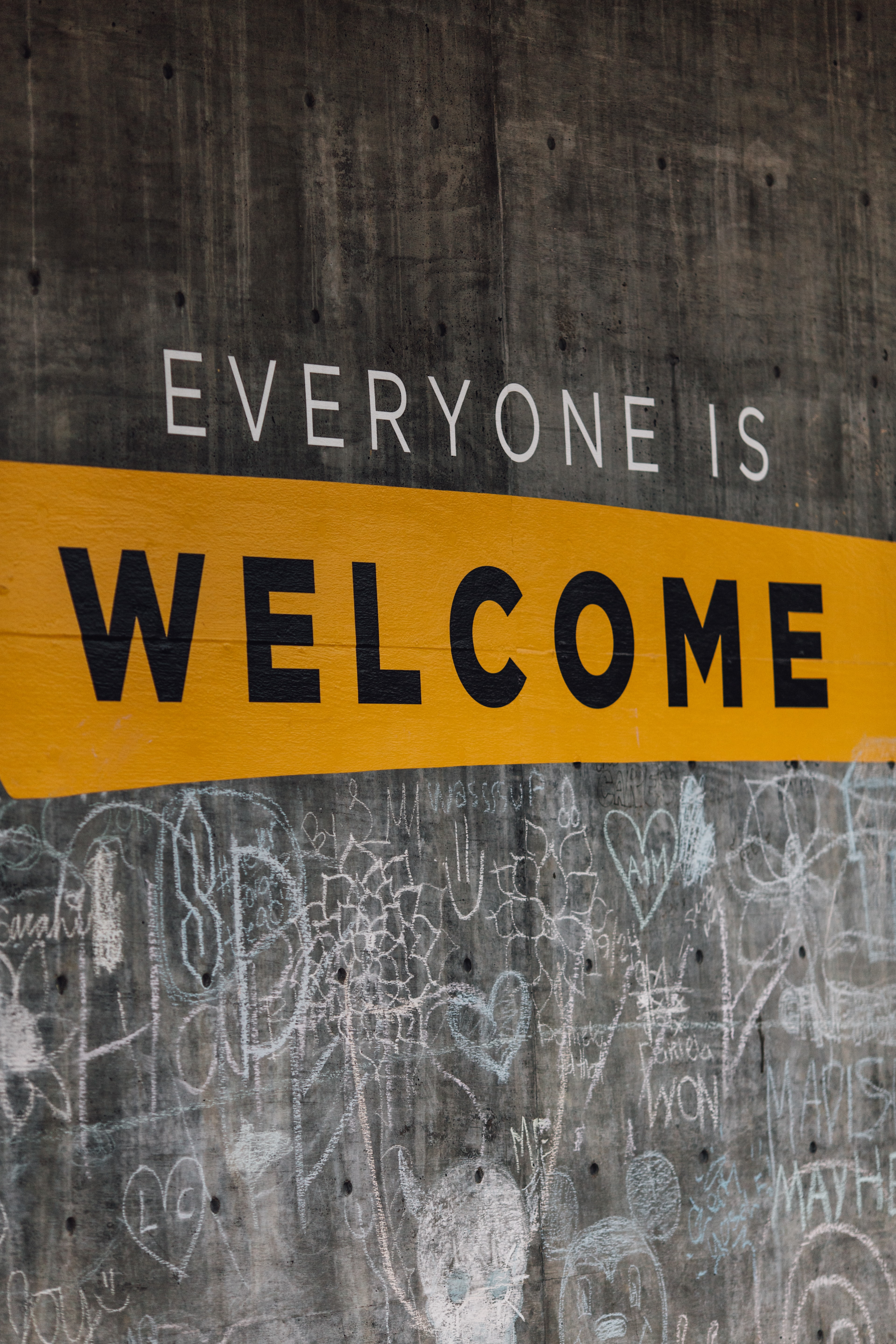 Sign on wooden wall reading “Everyone is welcome,” and chalk graffiti under sign